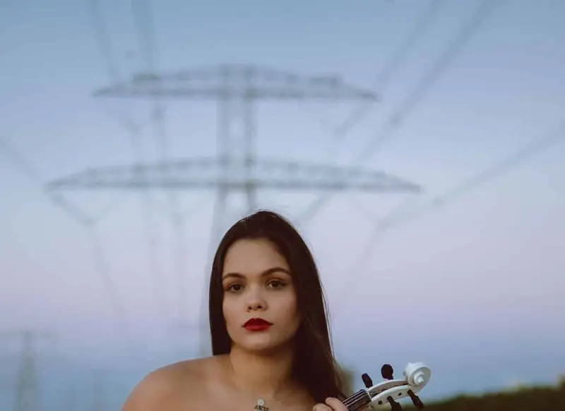 Female violinist in a black gown seated on sand holding a white electric violin